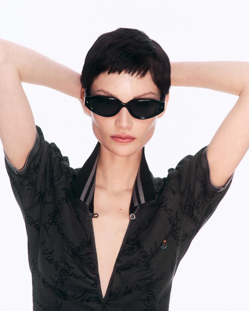 Woman wearing black sunglasses and dark patterned short-sleeved collared shirt poses with her hands behind her head on a white backdrop.