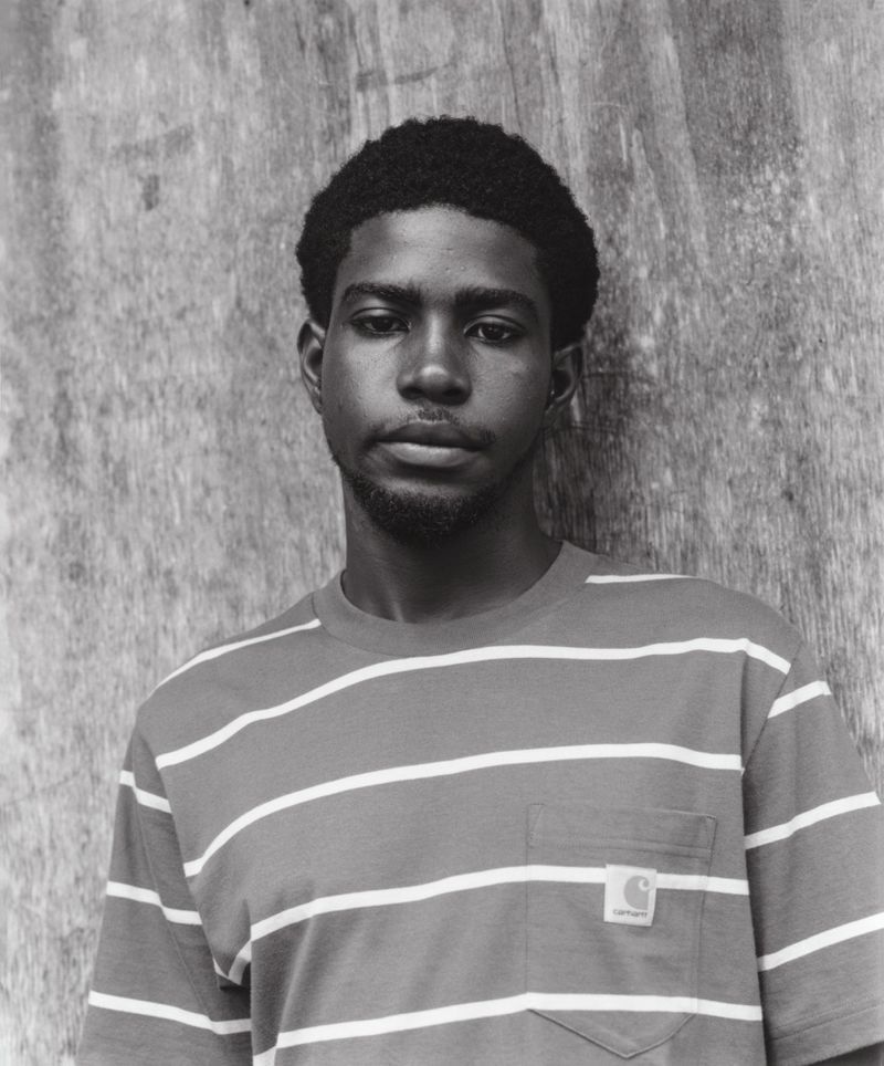 Monochrome portrait of a Black man in a striped t-shirt, gazing directly at the camera, set against a textured grey wooden background.