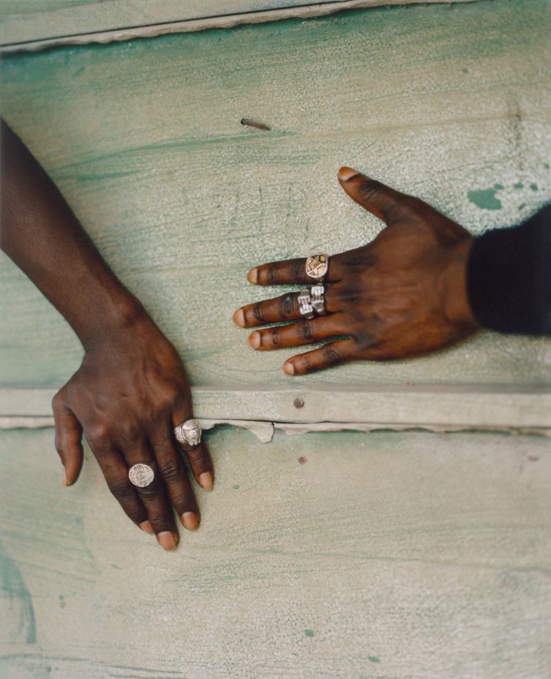 Close-up of two dark-skinned hands with jewelry, set against a textured green background, creating a striking contrast and focus on detail.