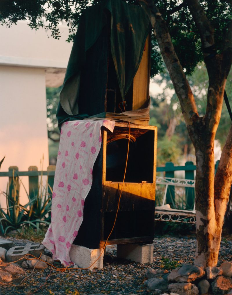 Open wooden box sits outside with a pink polka-dot cloth draped over it and a green tarp above; tree and greenery surround.