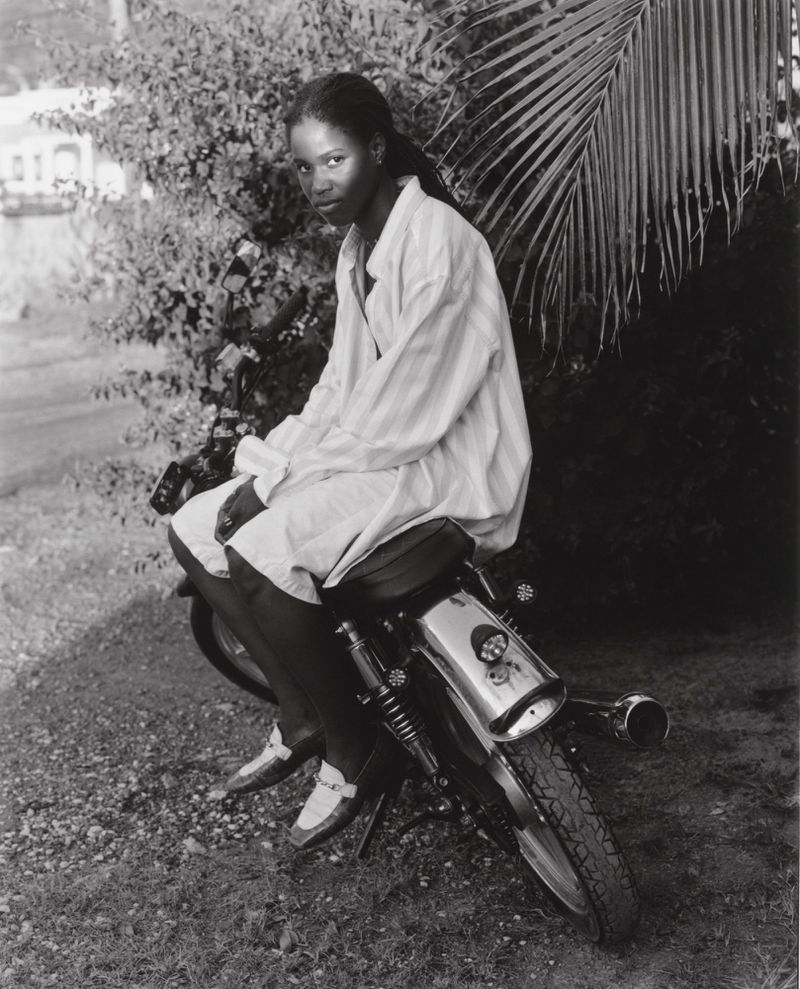 Black and white portrait of a Black woman seated on a motorcycle wearing a light-colored striped shirt and dress shoes in a garden setting.