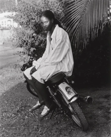 Black and white portrait of a Black woman seated on a motorcycle wearing a light-colored striped shirt and dress shoes in a garden setting.