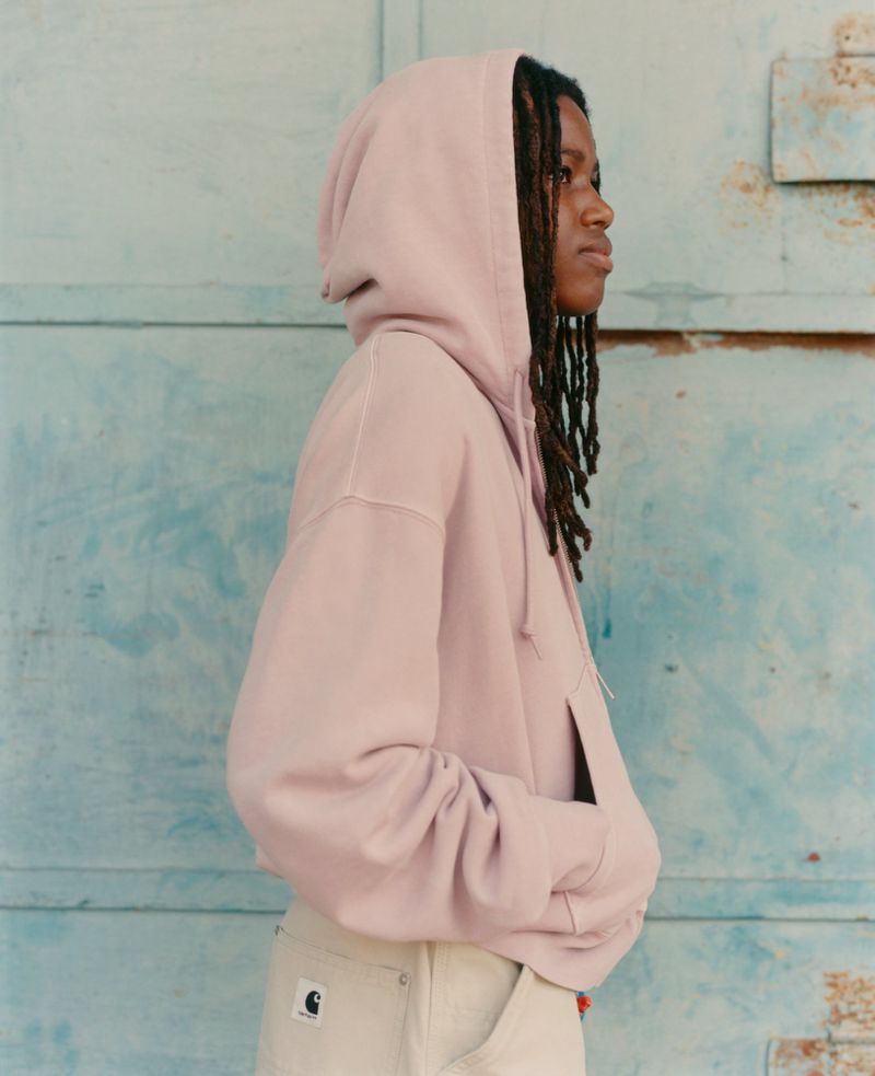 Profile of a young adult woman with dreadlocks wearing a pink hoodie and beige pants, posing against a weathered blue metal wall.