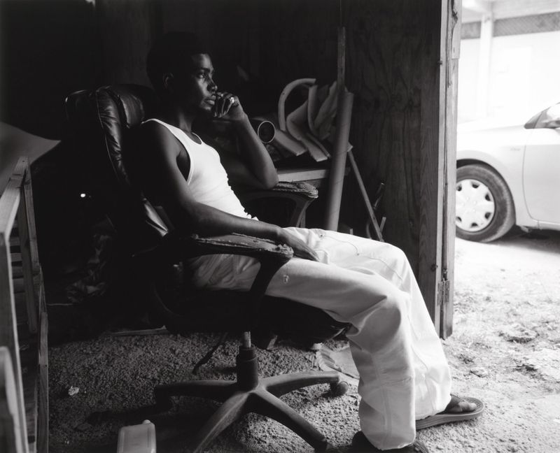 Black and white portrait of a man sitting in an office chair inside a room, wearing a tank top and pants, looking off to the side.