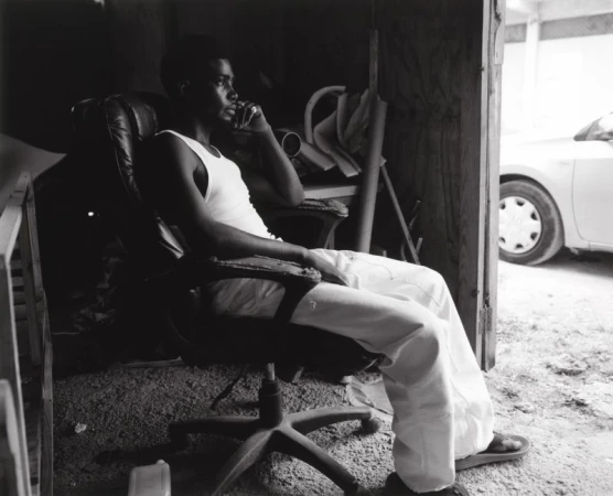 Black and white portrait of a man sitting in an office chair inside a room, wearing a tank top and pants, looking off to the side.