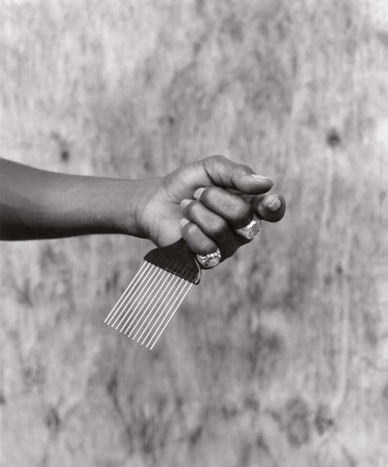 Close-up black and white shot of a dark-skinned hand holding a hair pick against a textured, neutral background, showcasing rings on the fingers.