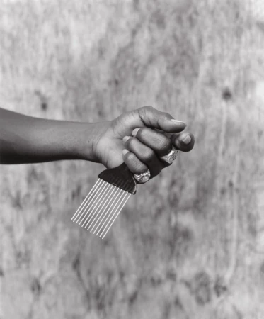 Close-up black and white shot of a dark-skinned hand holding a hair pick against a textured, neutral background, showcasing rings on the fingers.