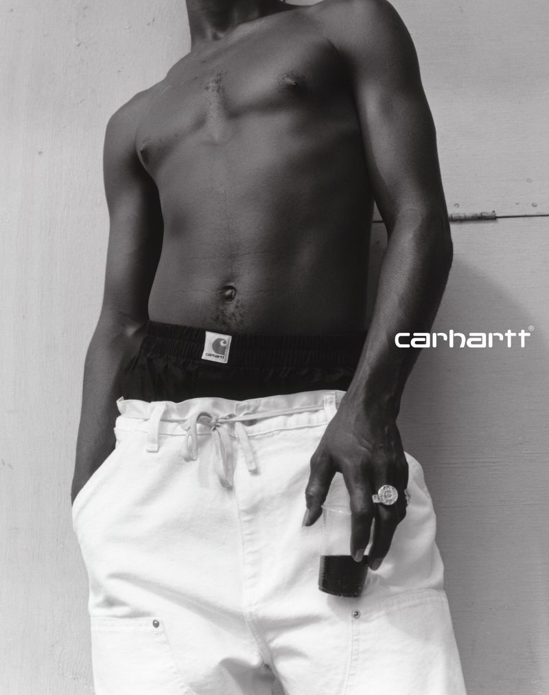 Monochromatic studio shot of a Black male torso, black pants, white trousers, silver ring, and black lighter against a wall.