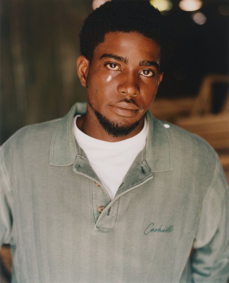 Portrait of a young adult African American man wearing a sage-green collared shirt with embroidered logo against a blurred background.