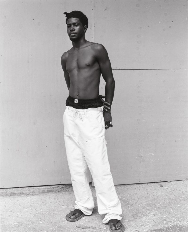 Monochrome portrait of a shirtless Black man in white pants and sandals, standing against a wall in neutral light, arms behind his back.