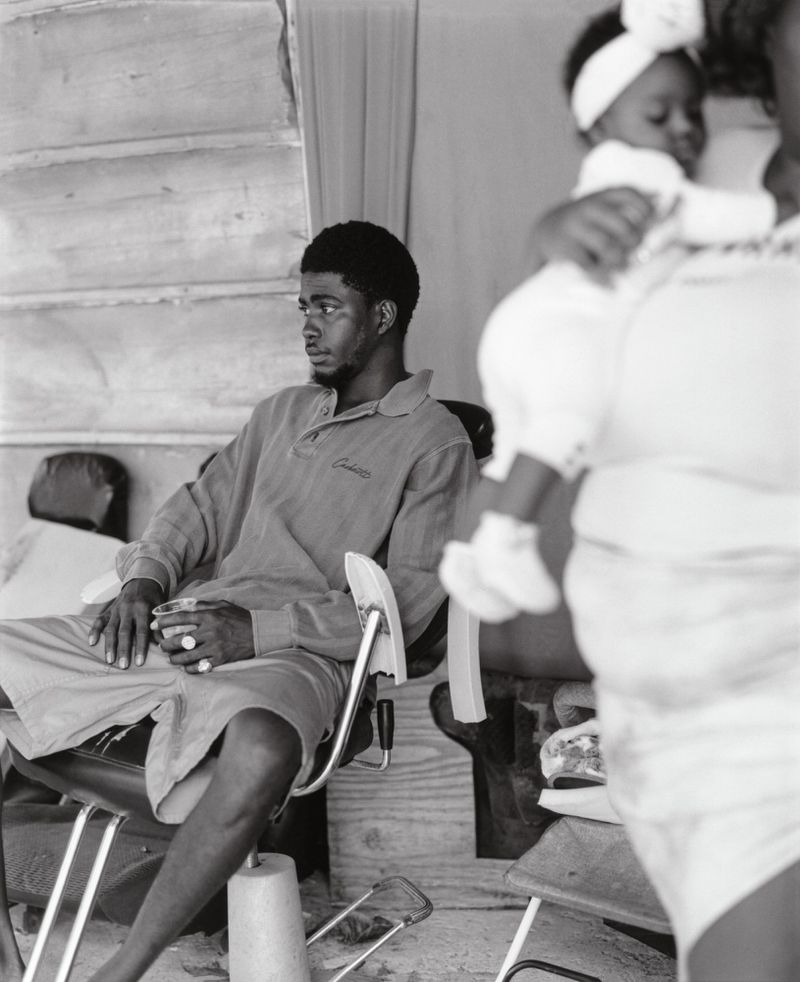 Black and white shot of an African-American man seated with an infant held by a woman partially visible in the background.