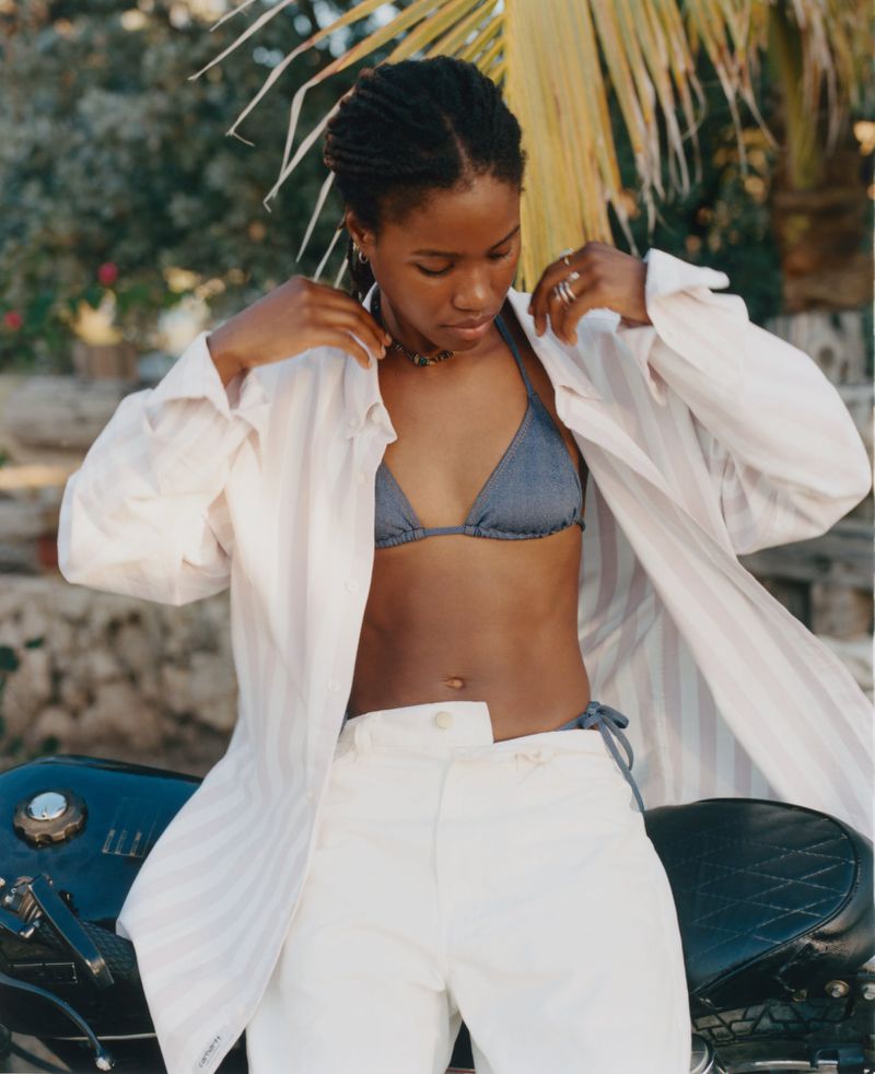 A woman in a bikini top, white pants, and a light coat poses near a motorcycle against a backdrop of palm trees and foliage.