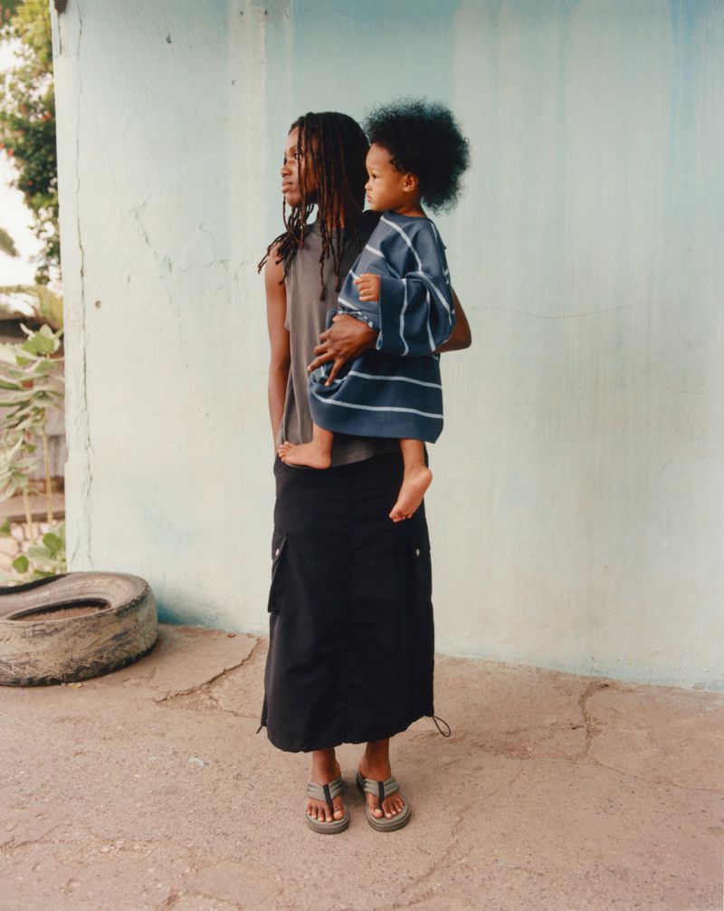 Woman holding a child outdoors against a light blue textured wall. Mother and child in casual clothing on a concrete surface.