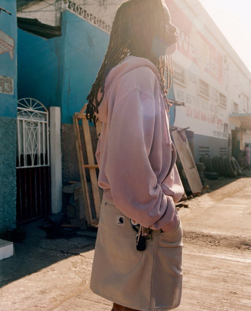 Adult female in mauve hoodie and khaki skirt stands on a sunlit urban street, looking up and to the right in a medium outdoor shot.