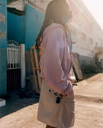 Adult female in mauve hoodie and khaki skirt stands on a sunlit urban street, looking up and to the right in a medium outdoor shot.