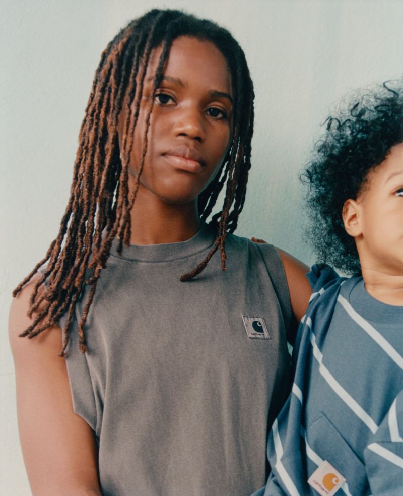 Two children pose indoors; one is wearing a gray Carhartt sleeveless top and the other a striped long-sleeved shirt, also by Carhartt.