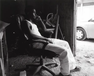 Black man wearing white tank top and trousers seated in an office chair inside a garage for Carhartt WIP S/S 25