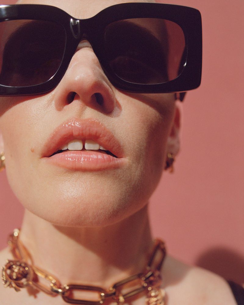 Close-up portrait of a woman with black sunglasses and a gold chain necklace against a pink background in an indoor setting.