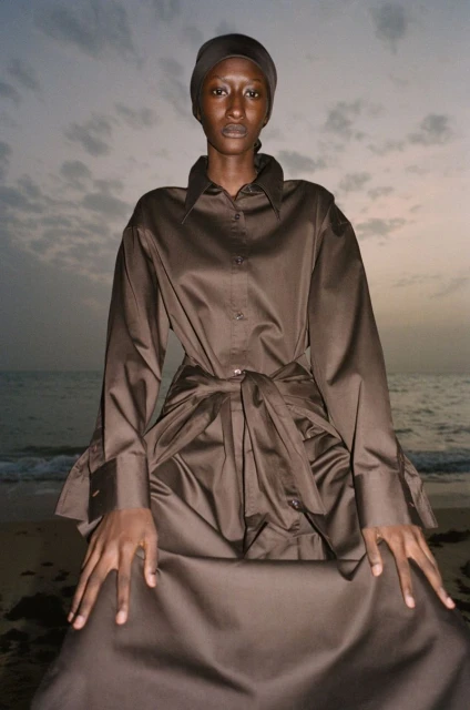 A dark-skinned woman in a brown dress and head covering poses on a beach with a cloudy sky and water in the background.