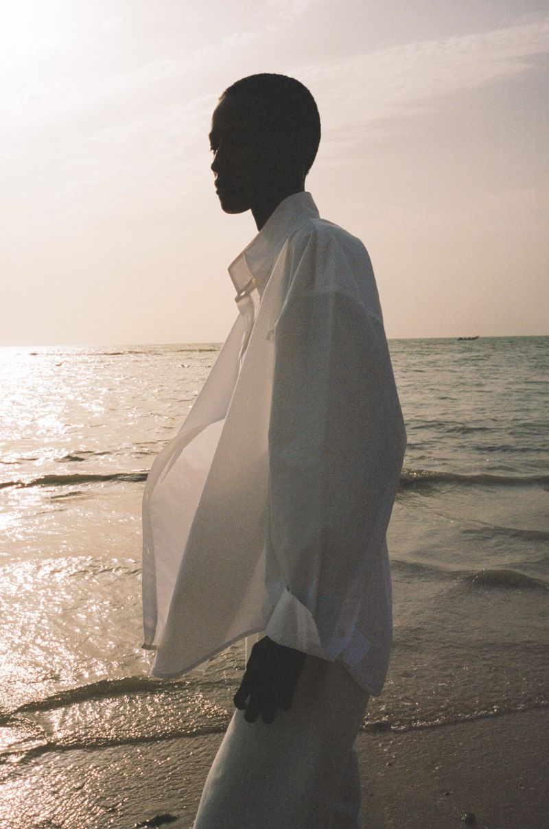 Model in white button down shirt standing on sandy beach at sunset for Teurn Spring 25 campaign
