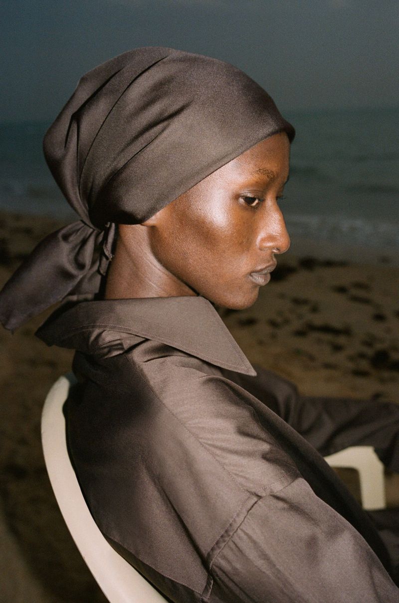 Woman in headscarf sitting on chair at beach. Close-up shot of African American female wearing brown head covering and shirt, ocean backdrop.