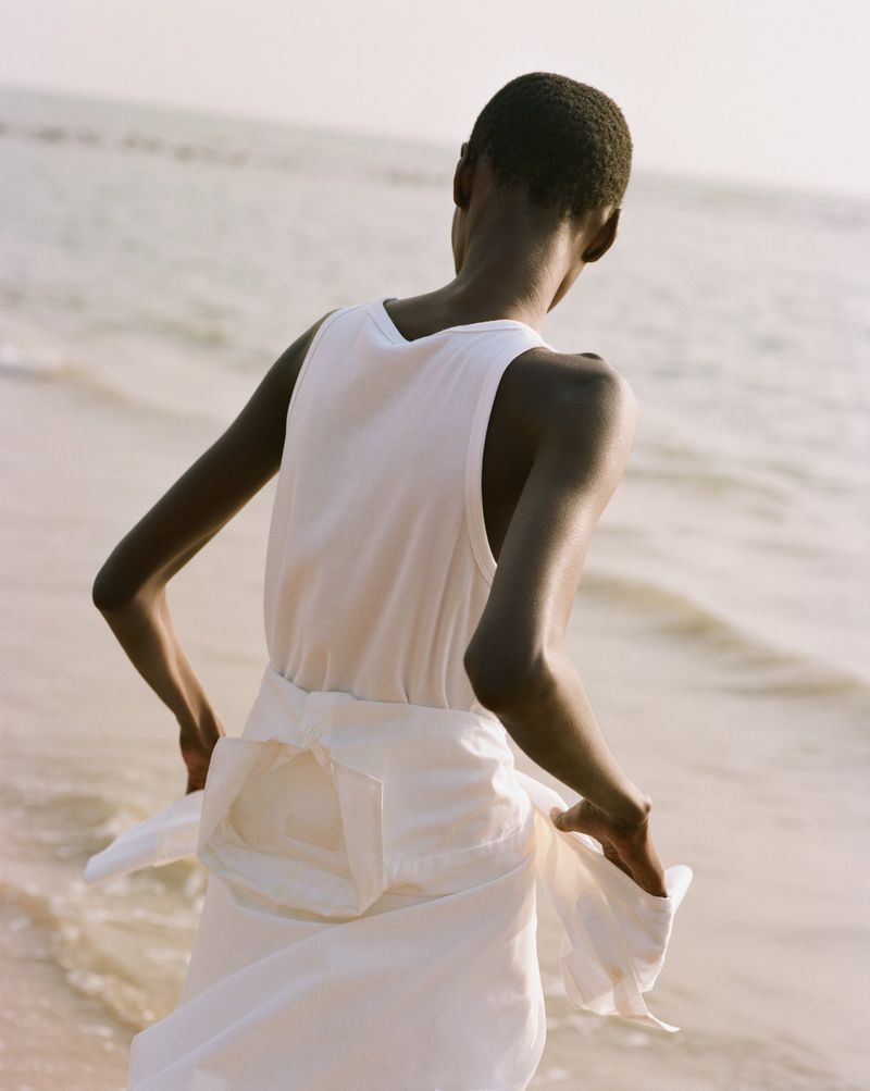A person stands at the beach at the water's edge, back to the camera, wearing a white top and white skirt, with the ocean in the background.