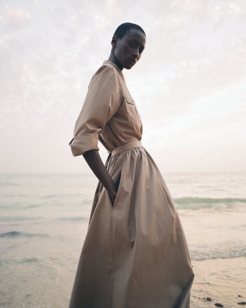 An adult woman with dark skin is standing on a beach in a long dress and button-up shirt, with the ocean and sky in the background.