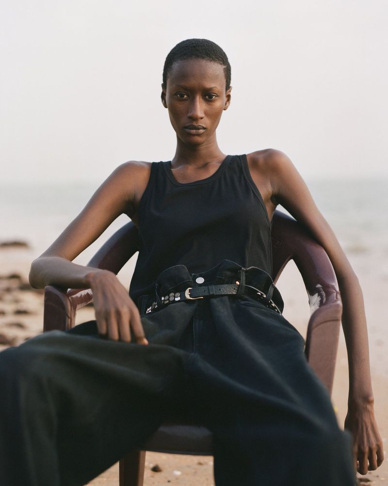 Elegant Black female model in tank top poses seated in chair on sandy beach with ocean backdrop. Beauty shot with natural light.