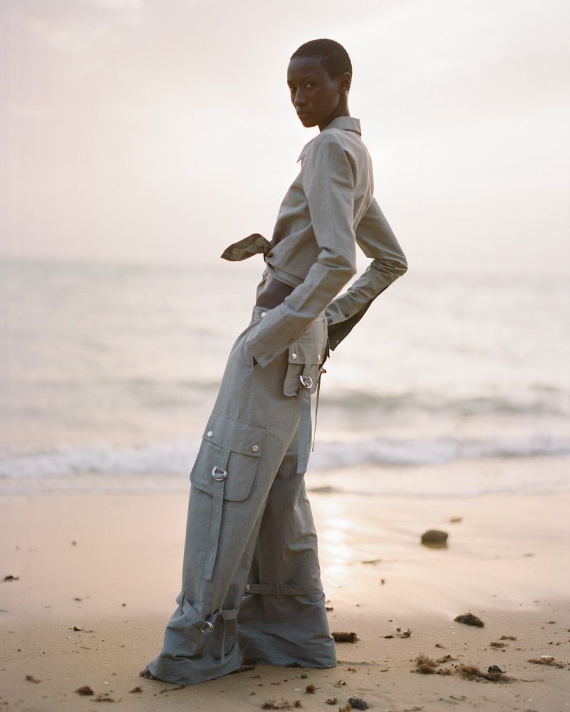 Woman poses on a sandy beach in a matching sage-green long-sleeve crop top and low-rise cargo pants near the ocean at dusk.