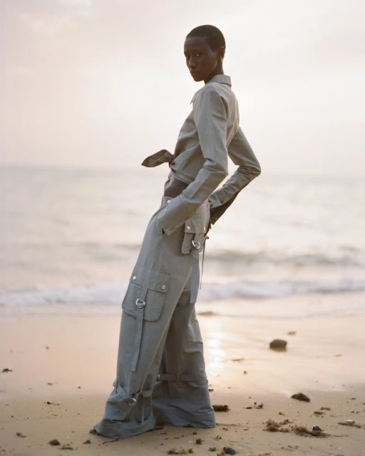 Woman poses on a sandy beach in a matching sage-green long-sleeve crop top and low-rise cargo pants near the ocean at dusk.