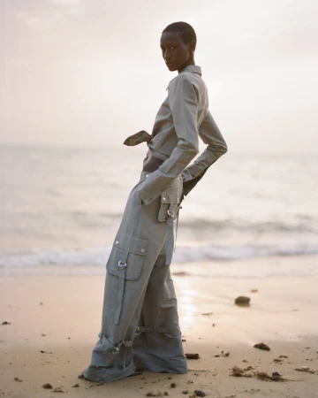 Woman poses on a sandy beach in a matching sage-green long-sleeve crop top and low-rise cargo pants near the ocean at dusk.