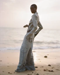 Model wearing a matching grey utility shirt and cargo trousers standing on a sandy beach at sunset.