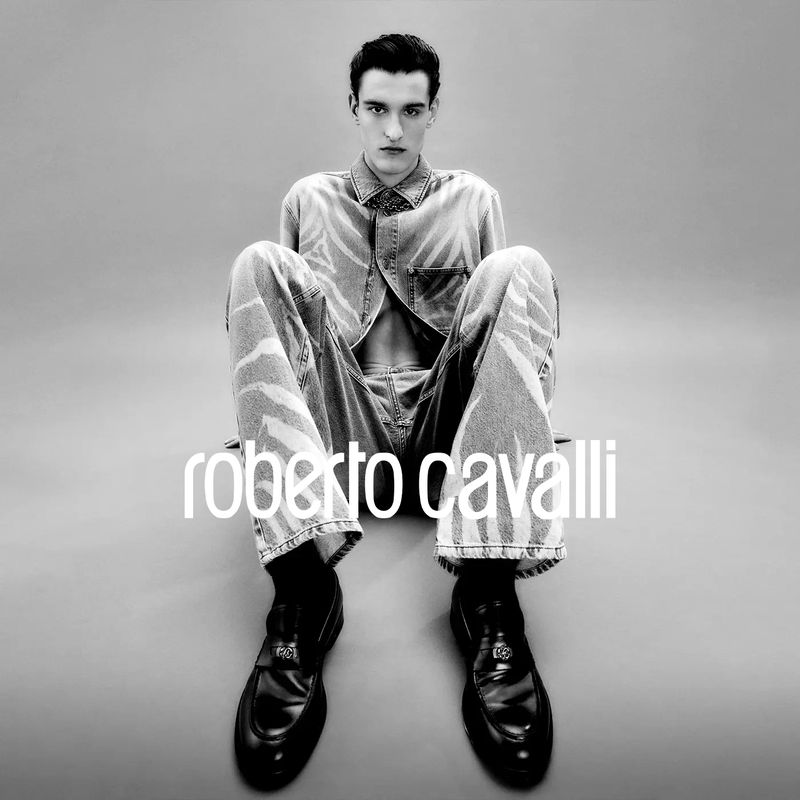 A young man with dark hair poses in an open denim shirt and matching denim pants with black loafers against a grey backdrop.