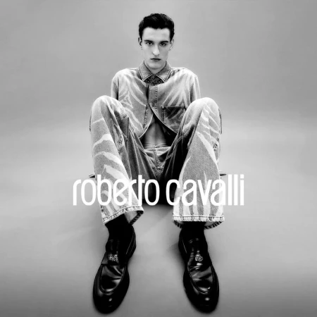 A young man with dark hair poses in an open denim shirt and matching denim pants with black loafers against a grey backdrop.