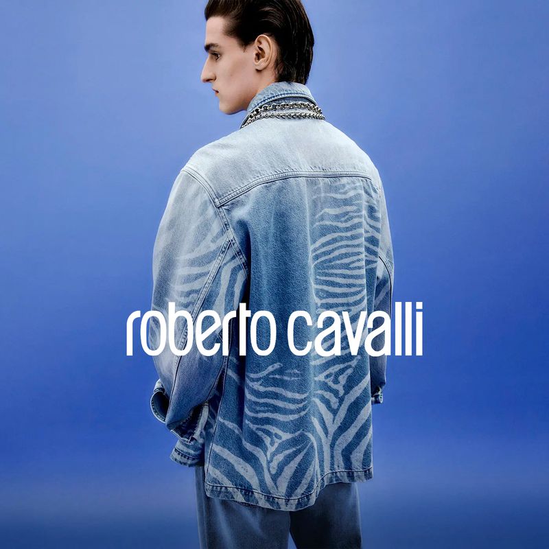 Fashion shot featuring a male model wearing a denim jacket with zebra print. He's posing against a blue background with brand logo.