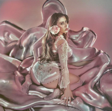Portrait of a woman kneeling on shiny pink fabric, wearing lace lingerie and a long-sleeved top, with a flower in her hair.