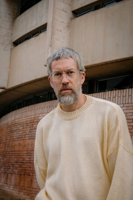 Portrait of an adult man with a beard and glasses, wearing a cream sweater, standing in front of a modern concrete building.