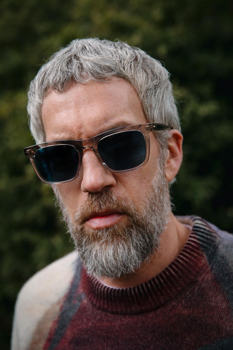 Adult male with gray hair and beard wearing sunglasses, looking at the camera, against a blurred green background. Close-up shot.