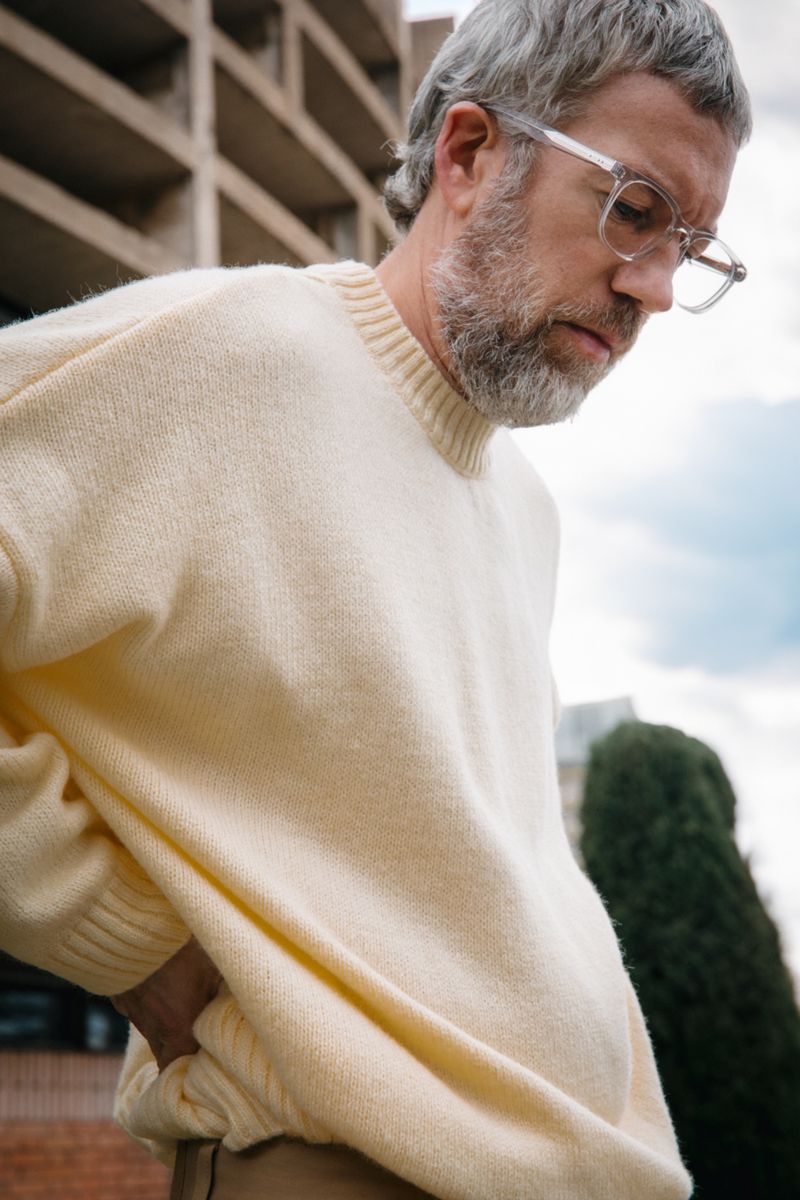 Man wearing clear-framed eyeglasses and a cream knit sweater against an outdoor architectural background.