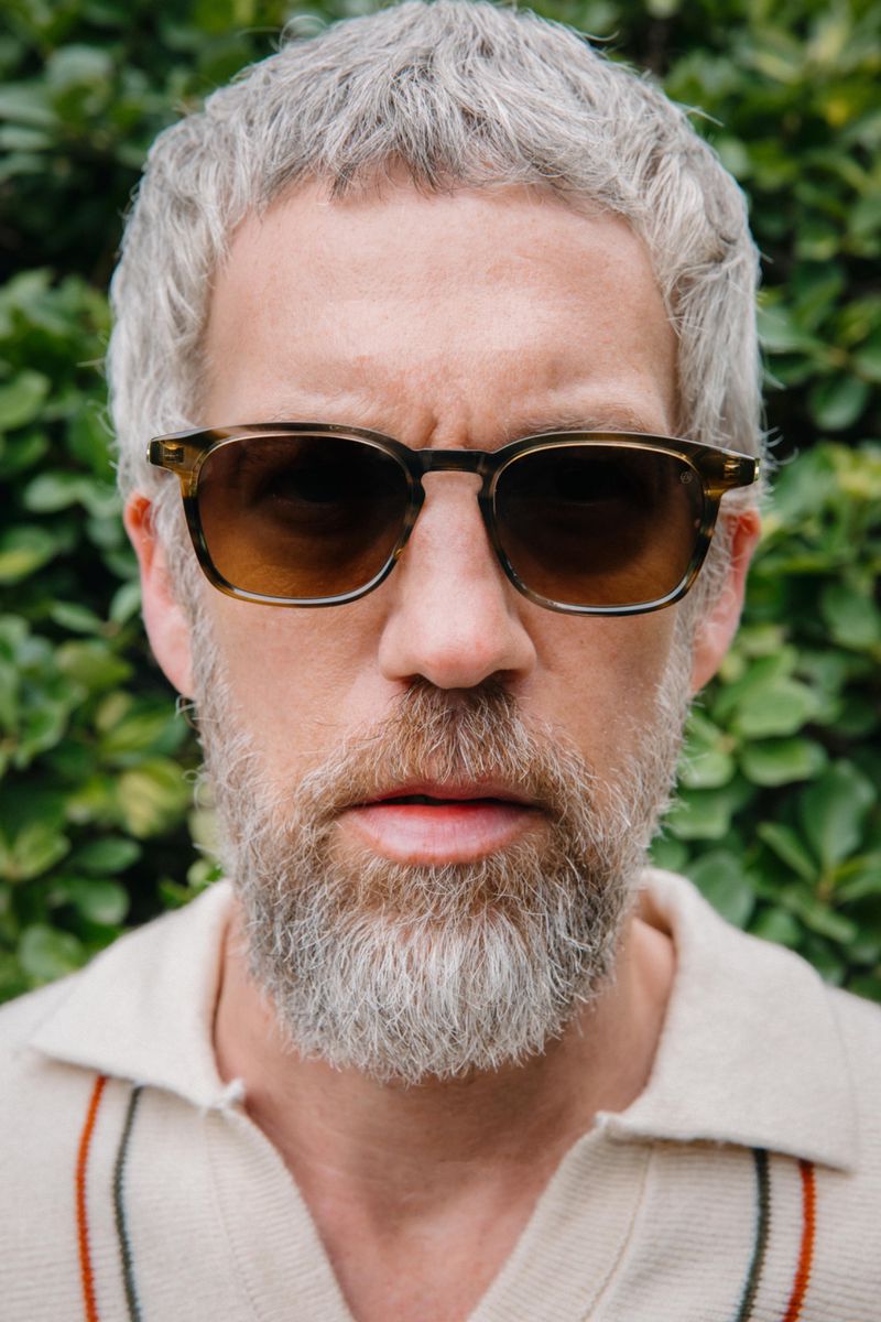 Headshot of a man with grey hair and a beard wearing sunglasses, looking directly at the camera against a green background.