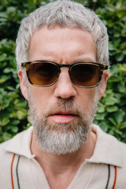 Headshot of a man with grey hair and a beard wearing sunglasses, looking directly at the camera against a green background.