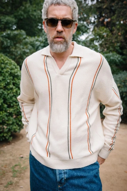 Adult male wearing a cream-colored sweater with blue jeans stands outdoors among green foliage on a sunny day.