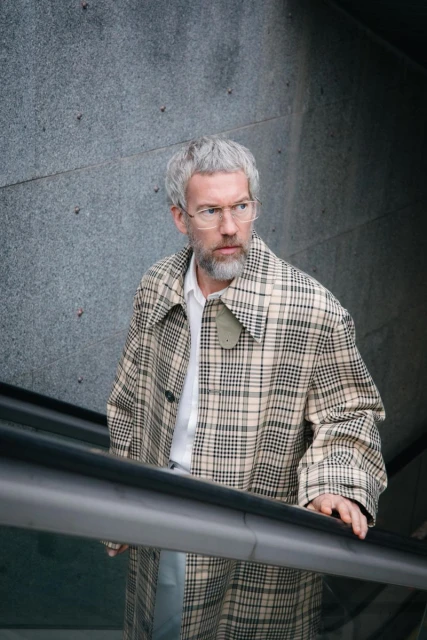Man with grey hair and beard in plaid coat on escalator looks up, set against a textured grey wall. Eye-level, medium shot.