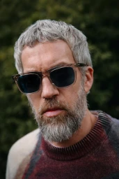 Close-up portrait of a man wearing clear-framed sunglasses and a patterned sweater outdoors against green foliage.
