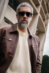 Man wearing sunglasses and brown leather jacket with cream sweater against a textured concrete architectural exterior
