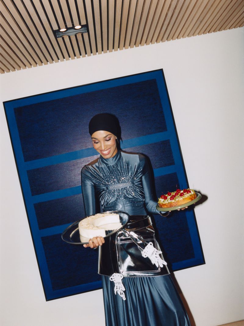 A woman wearing a dark blue dress and head covering smiles as she holds trays with cake, posing in an interior with modern art.