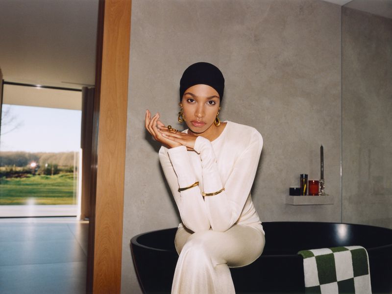 Elegant woman in white outfit and black head covering sits in a modern bathroom with a dark tub and view to a green landscape.