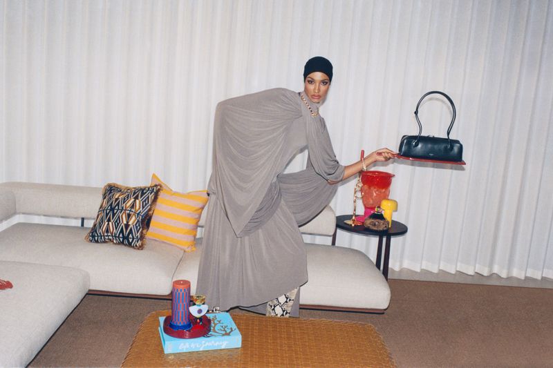 A woman in a grey dress and black head covering poses in a room with a couch, table with colorful objects, and holds a black handbag.