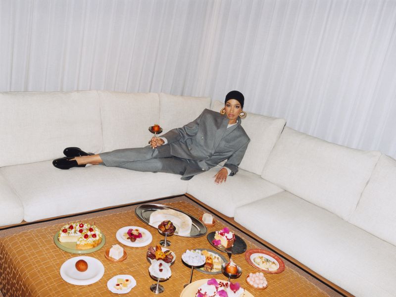 Woman in a grey suit lounges on a white sofa with desserts arranged on a low table; the scene is lit with soft, natural light.