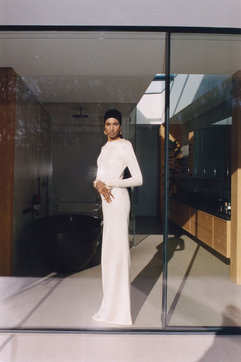 A woman in a long white dress stands in front of a modern bathroom, illuminated by natural light streaming through large glass doors.